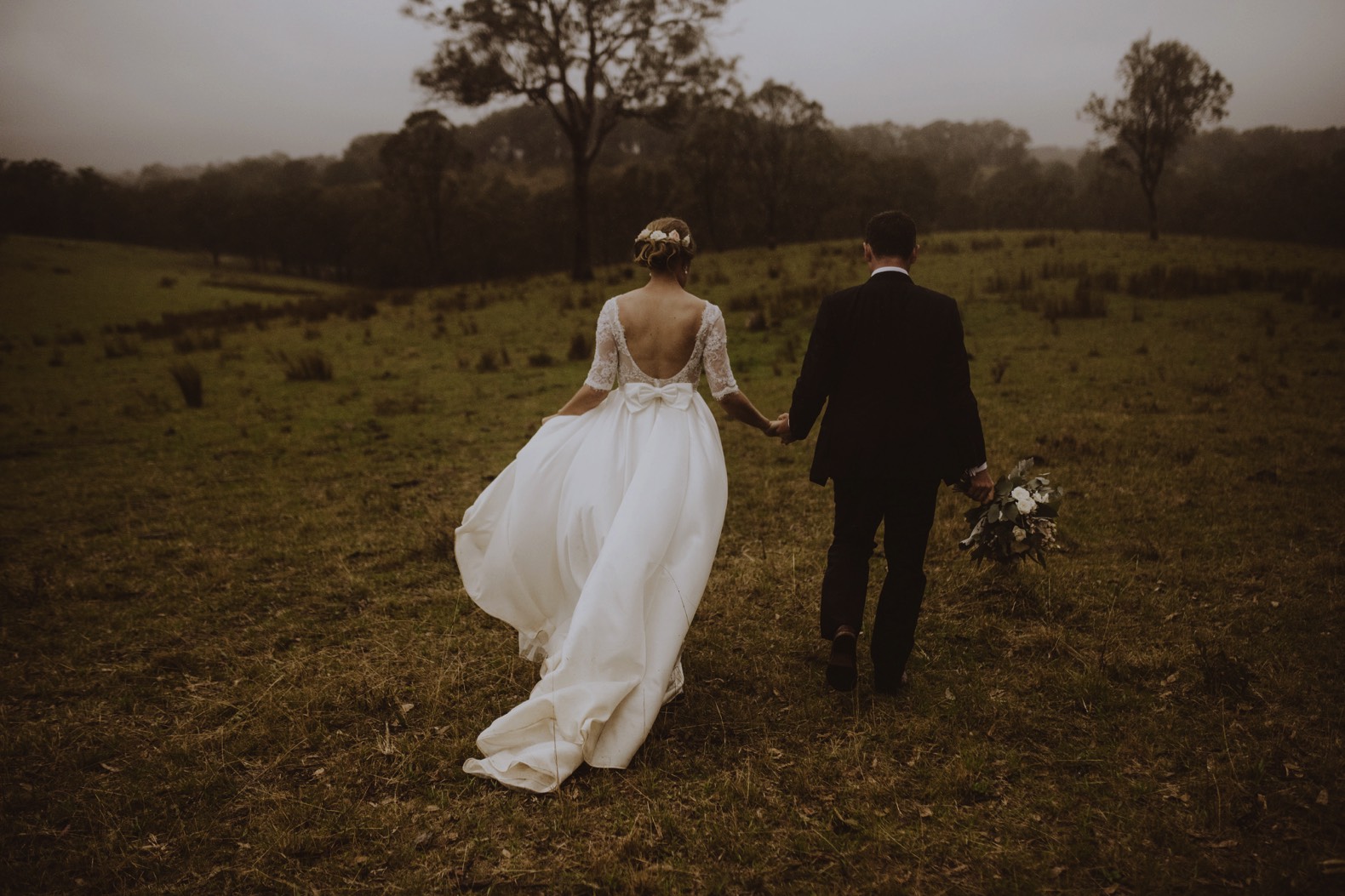 photo of bride and groom walking away hand in hand in a field in the rain